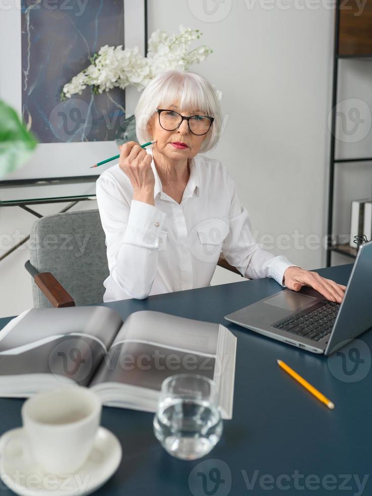 trött senior vacker grått hår kvinna i vit blus läser dokument på kontoret. arbete, seniorer, frågor, hitta en lösning, uppleva koncept foto