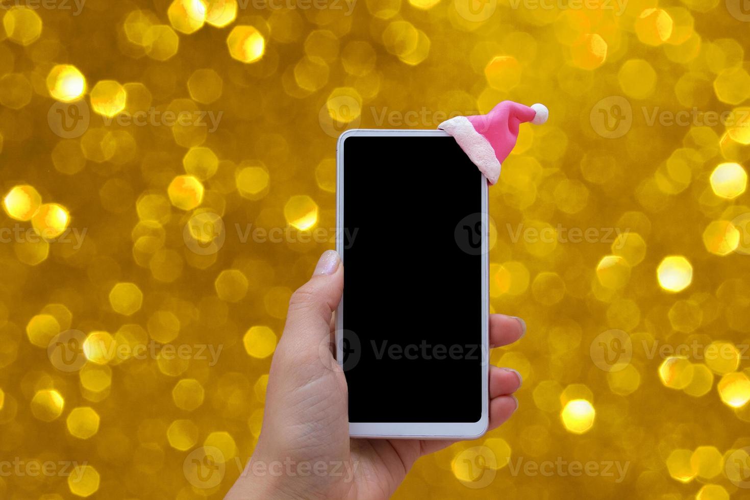 mock up telefon med tomtehatt i kvinna hand på gyllene bakgrund med boke. jul online försäljning koncept foto