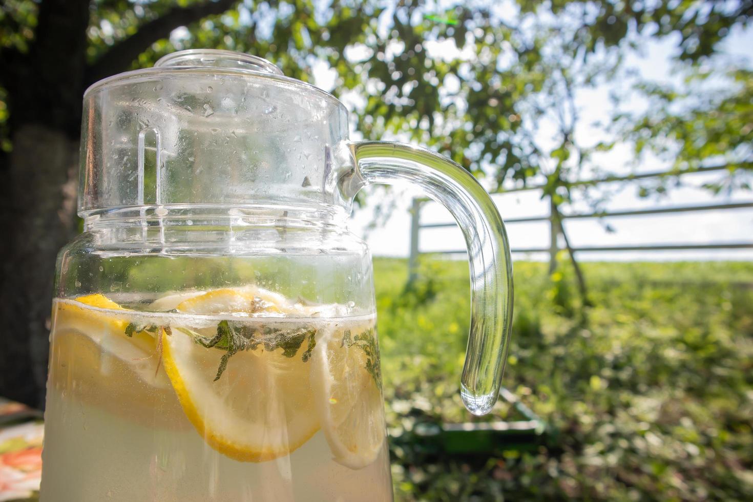 hemgjord lemonad gjord av citroner i en stor glaskanna på bordet i trädgården. en kanna med citron och mynta står på gatan mot bakgrund av grönska en varm sommardag. foto