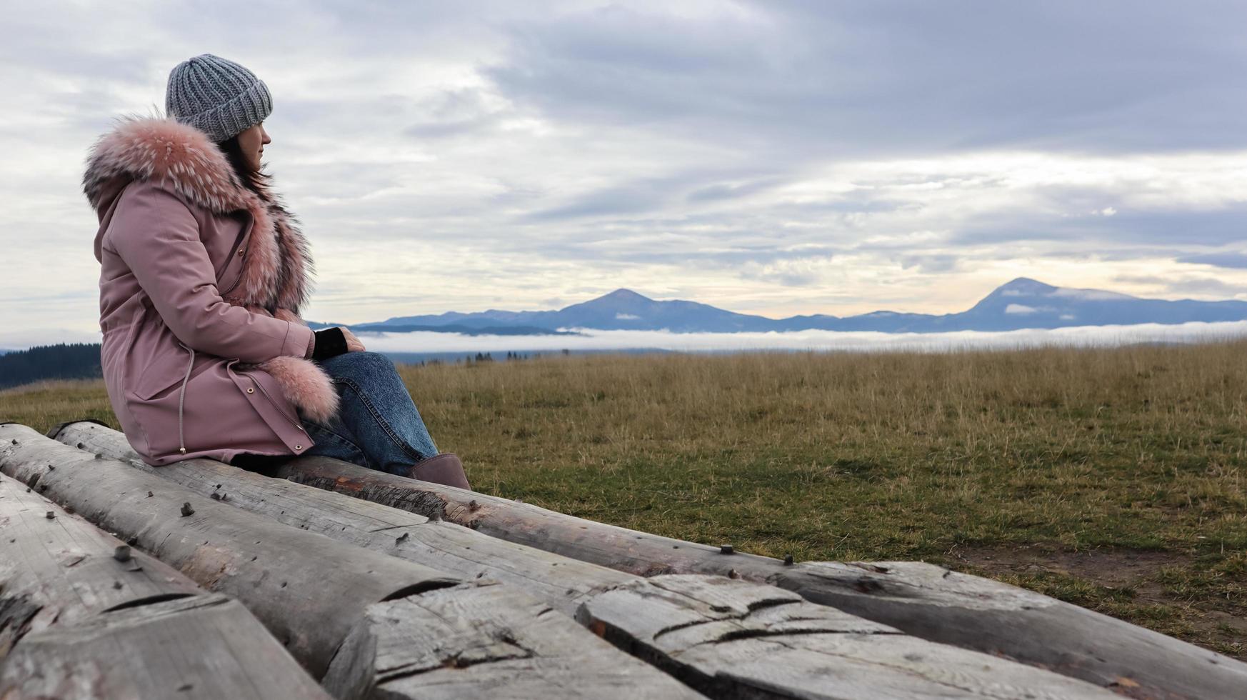 en kvinna sitter med ryggen på trästockar högt uppe i bergen i kallt höstväder och njuter av den vackra utsikten över det bergiga naturlandskapet. foto