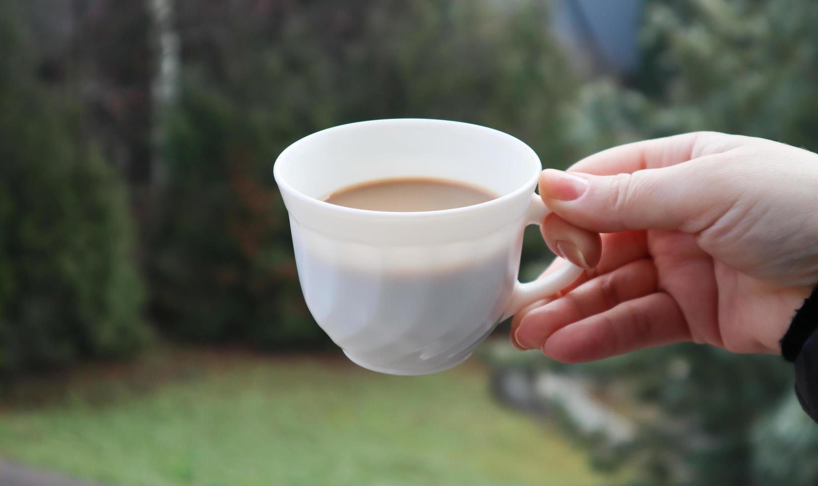 flickans hand håller en kopp vitt kaffe och har en suddig grön naturbakgrund. mugg i en kvinnlig hand på en bakgrund av berg. foto