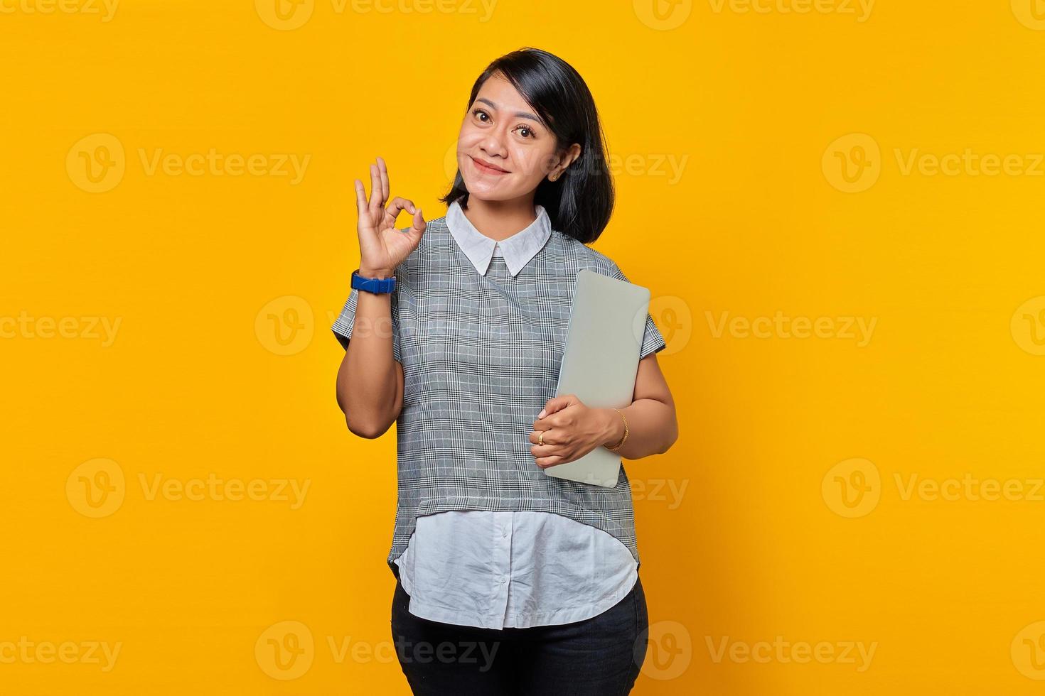 vacker asiatisk kvinna med laptop ler och visar okej tecken över gul bakgrund foto