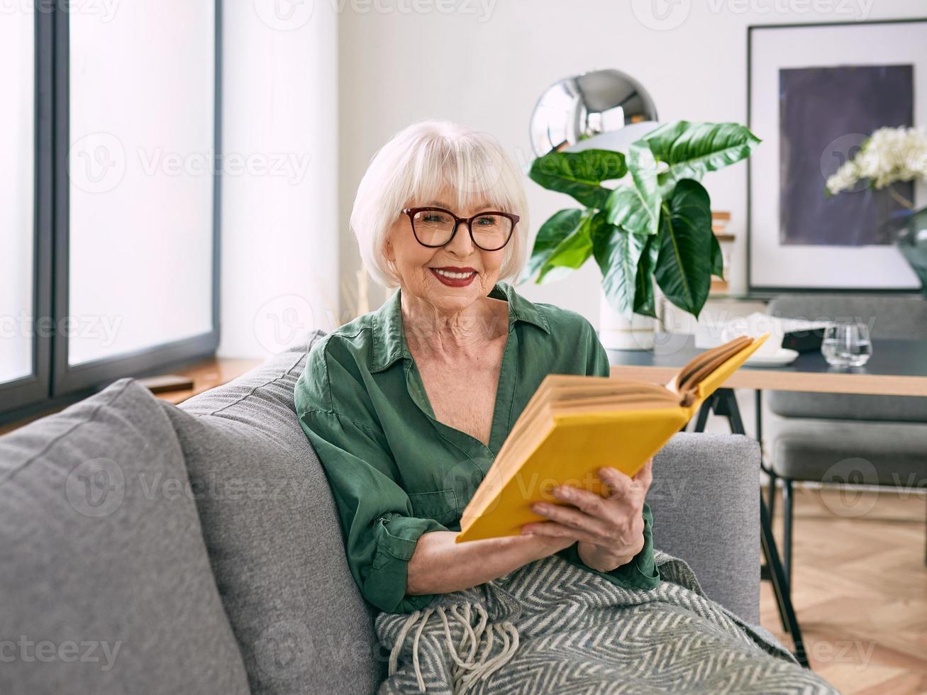 glad senior kvinna sitter i soffan och läser en bok hemma. utbildning, mogna, fritid koncept foto