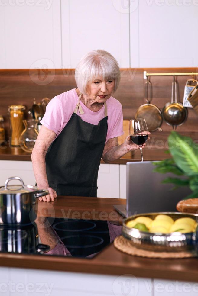 senior glad kvinna lagar mat på modernt kök med bärbar dator. mat, utbildning, livsstilskoncept foto