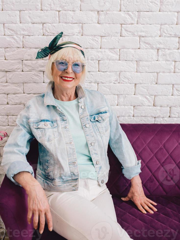 äldre senior snygg kvinna i blå solglasögon och jeansjacka sitter på soffan i elegant loftinteriör i salongen. stil, mode, vänta, anti ålder koncept foto