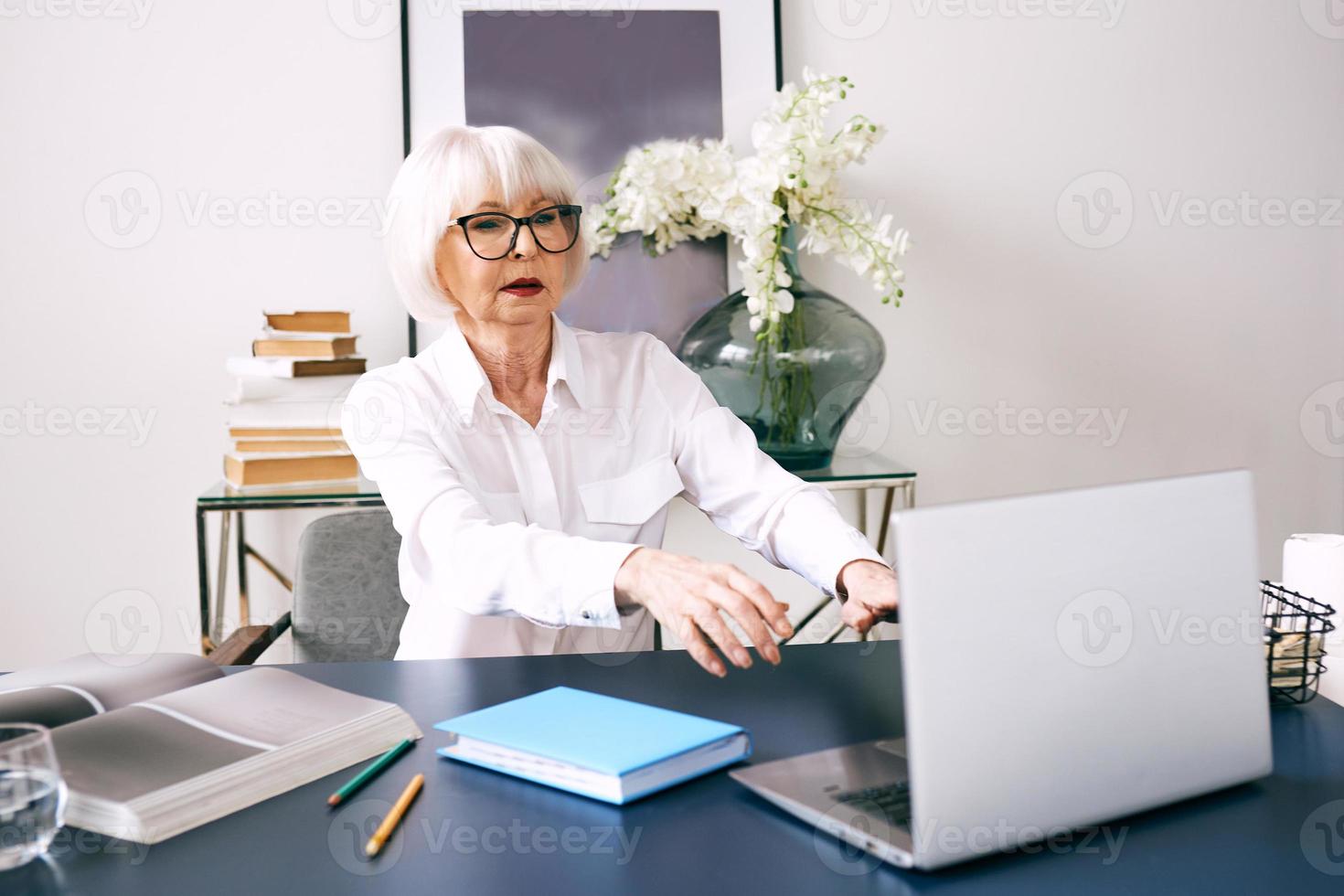 trött senior vacker grått hår kvinna i vit blus som arbetar på laptop på kontoret. arbete, seniorer, frågor, hitta en lösning, uppleva koncept foto