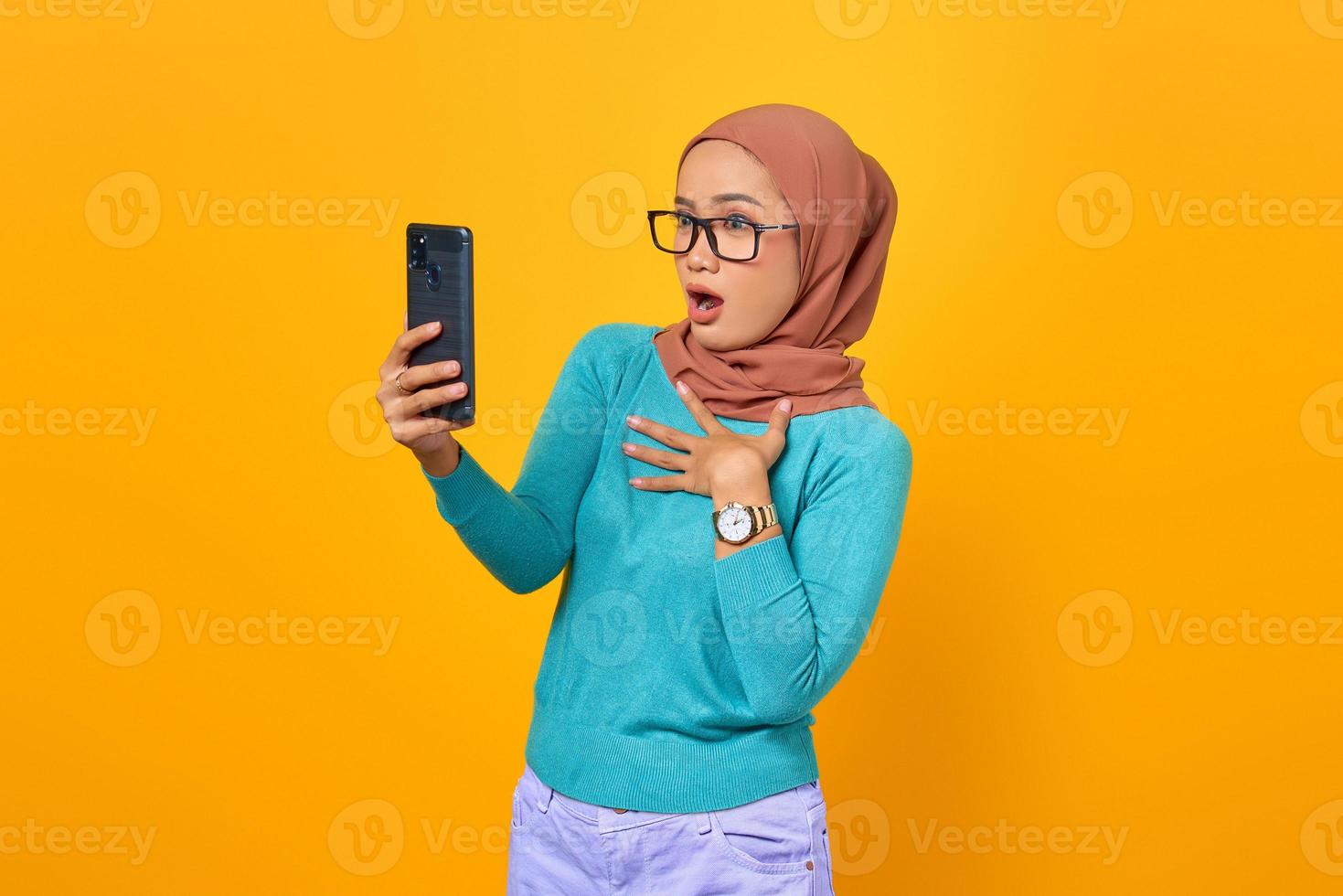 chockad vacker ung asiatisk kvinna tittar på smartphone-skärmen isolerad på gul bakgrund foto