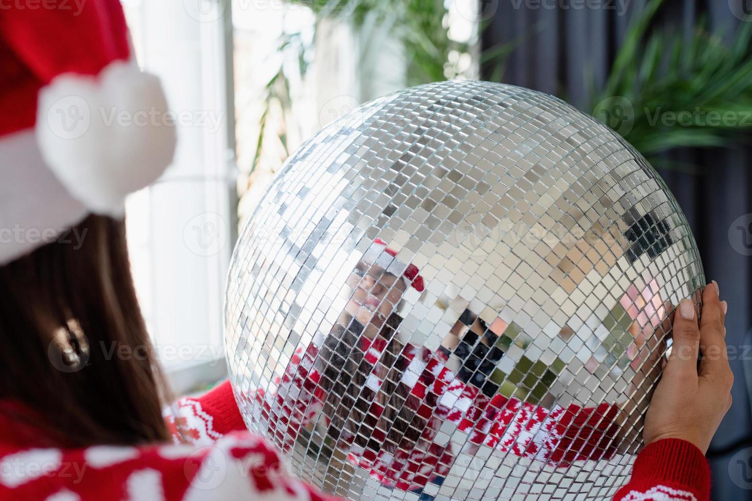 ung kvinna i tomtehatt håller spegeln disco boll stående vid julgranen foto