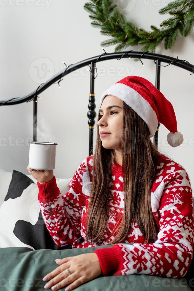 ung kvinna i röd tröja och tomtehatt sitter på sängen och firar jul med en kopp varm dryck foto
