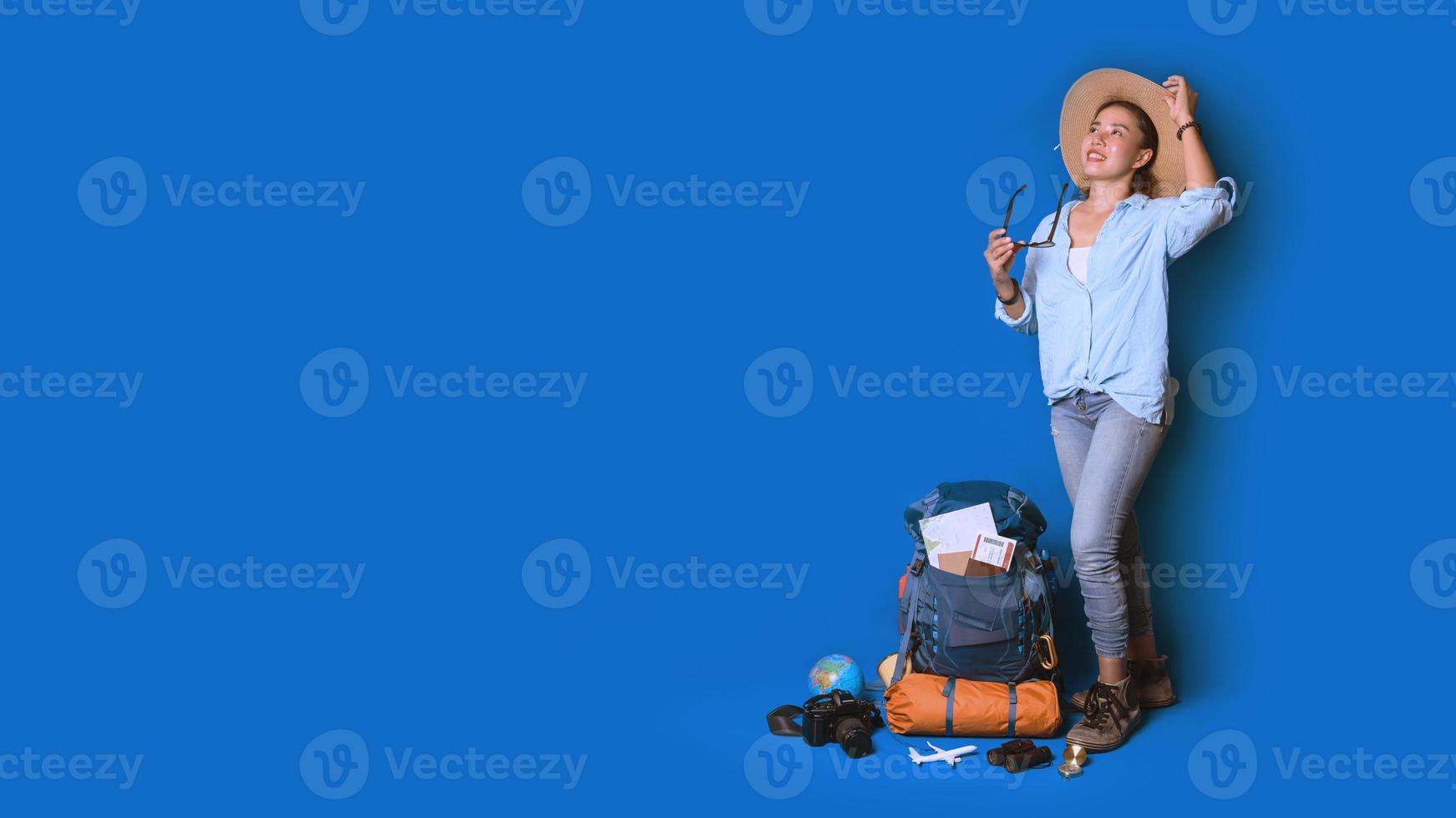 ung asiatisk resenär glad kvinna i blå skjorta med ryggsäck med och utrustning för resenärer semester med en karta, på blå färg bakgrund. reseryggsäck foto