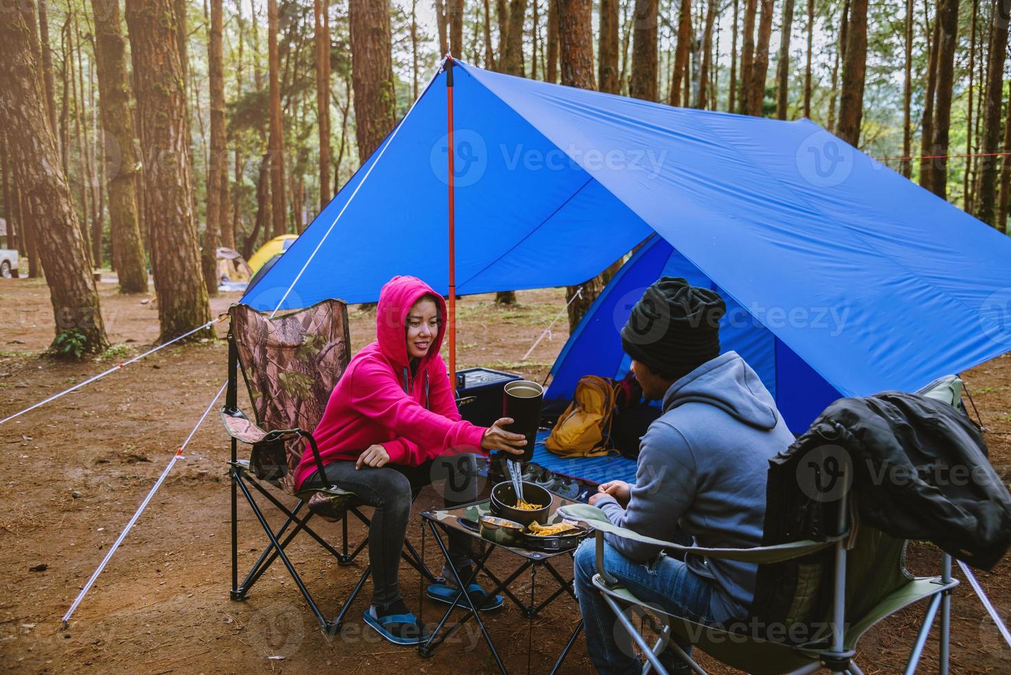 ungt asiatiskt par glada njuter av camping i tallskogen sitter och äter mat på campingsidan mitt i naturen. foto