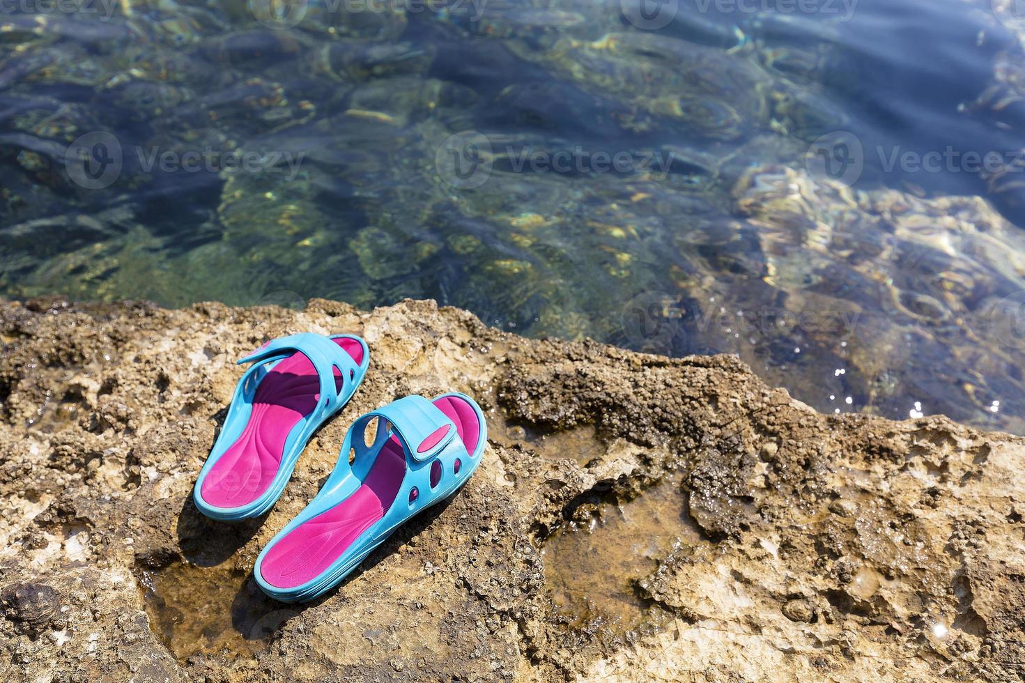 strandtofflor ligger på en sten nära det genomskinliga havet foto