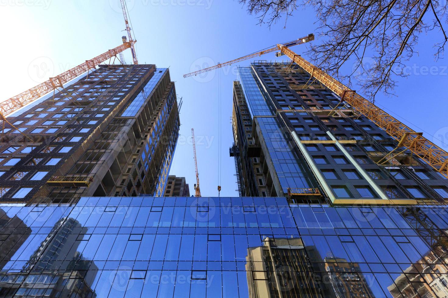 glasfasaden, en reflektion av den blå himlen och kranar nära en modern betongbyggnad under uppförande. foto
