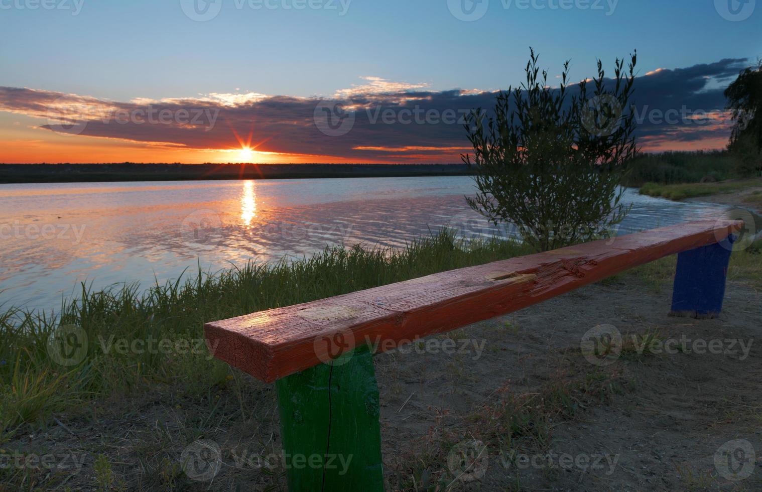 majestätisk gyllene gryning med en kontur av träd och en gammal träbänk på flodstranden foto