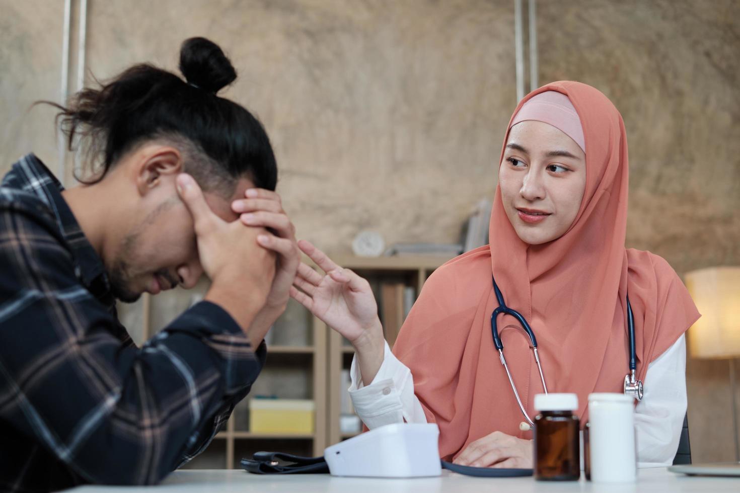 en ung vacker läkare, en person som är kvinnlig muslim uppmuntrar och tröstar manlig patient av asiatisk etnicitet som är ledsen och sjuk, på hälso- och sjukvårdsklinikens kontor på sjukhus. foto