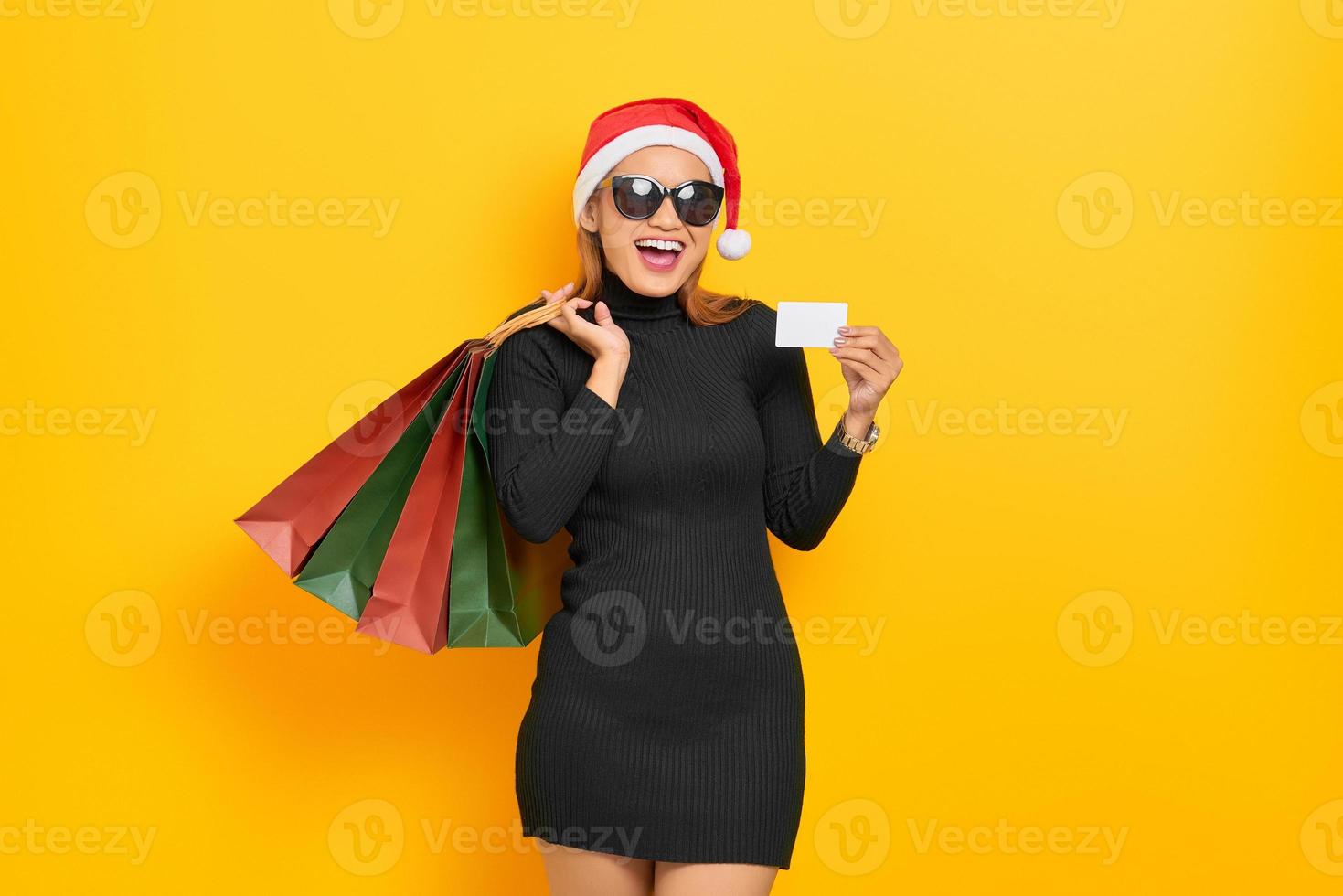 leende ung asiatisk kvinna i jultomtehatt och solglasögon som håller shoppingkassar och plastkort isolerade över gul bakgrund foto