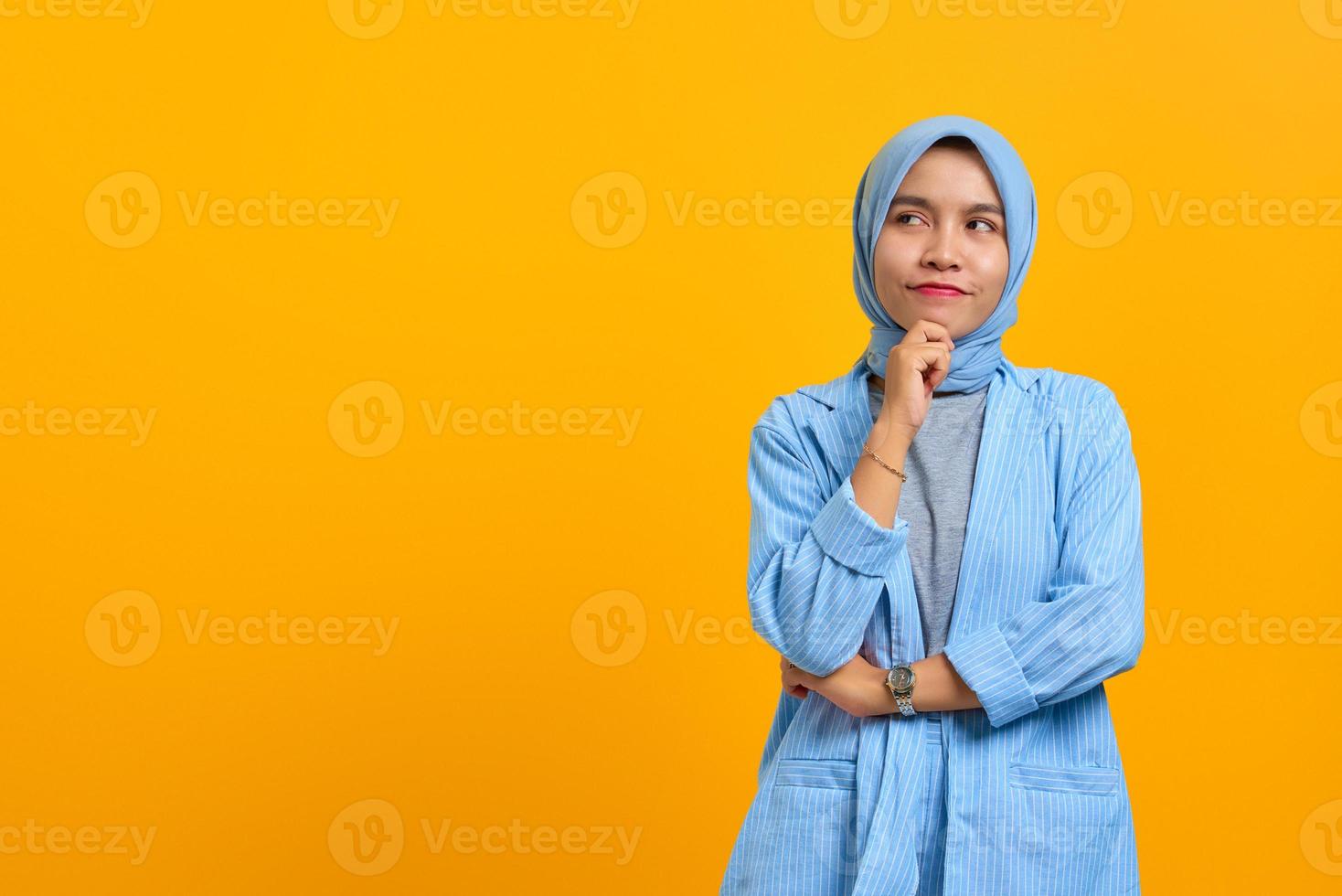 fundersam ung asiatisk kvinna tänker på fråga med händerna på hakan över gul bakgrund foto