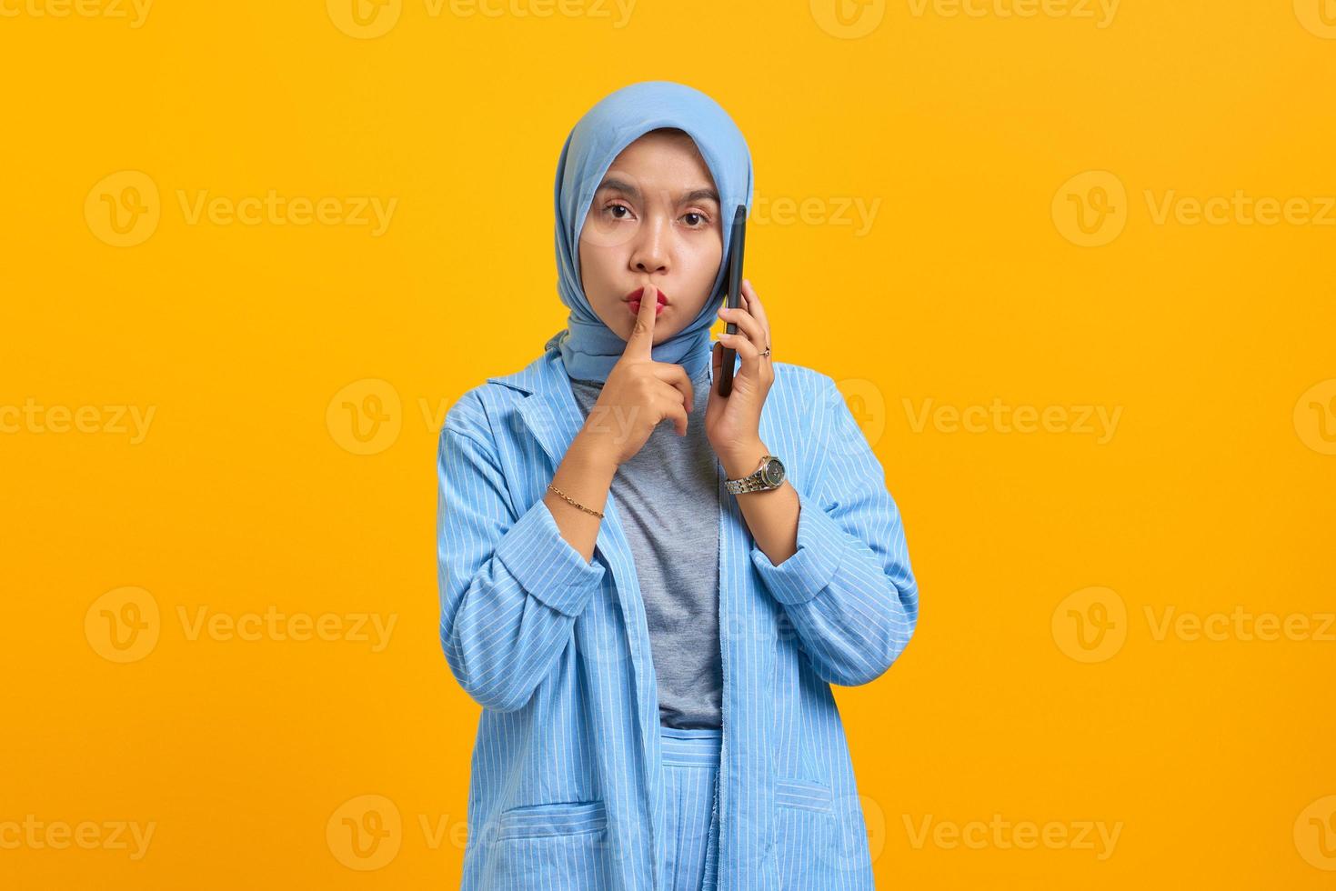 ung asiatisk kvinna pratar i mobiltelefon och visar tystnadsgest över gul bakgrund foto