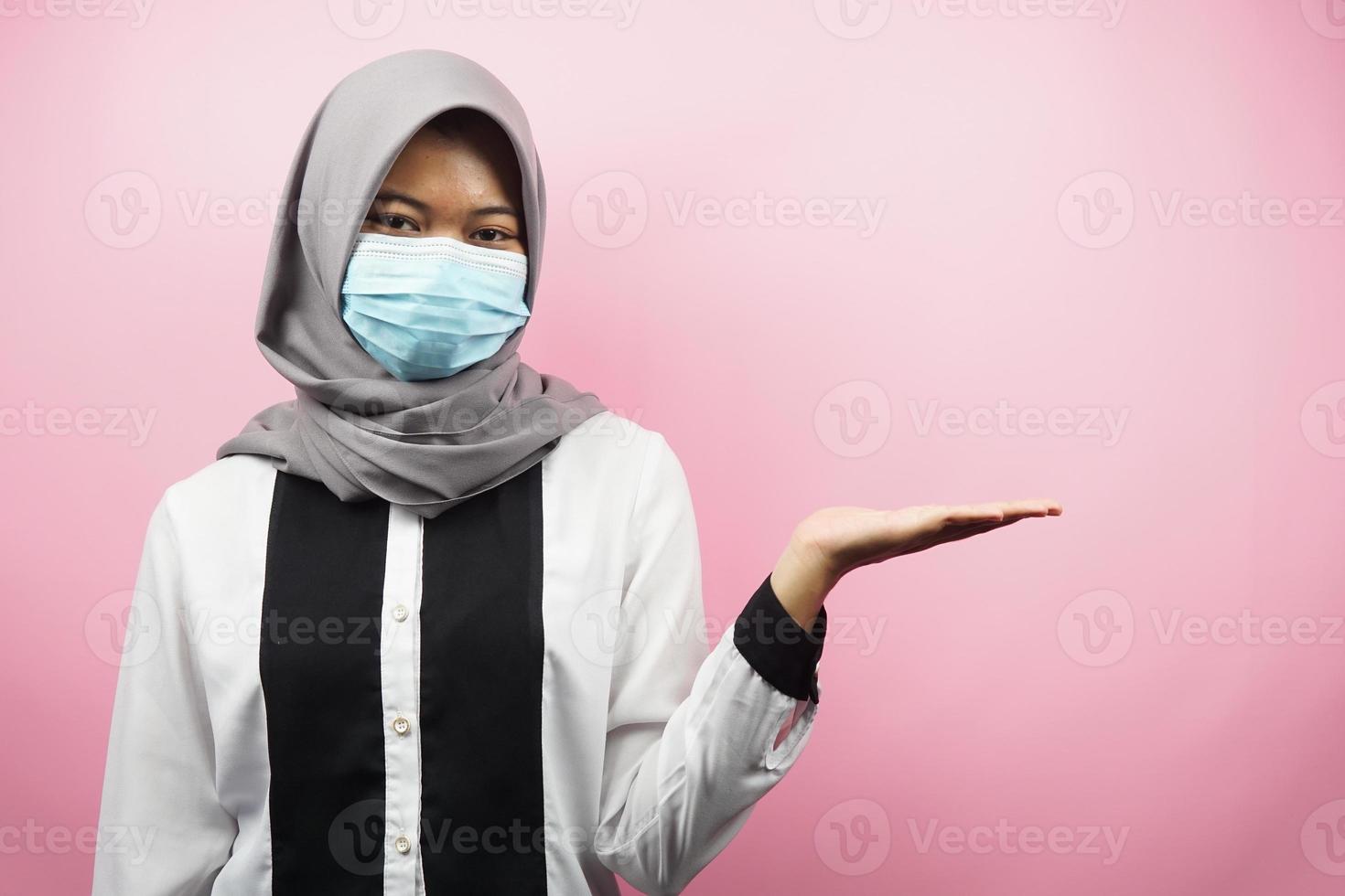 muslimsk kvinna som bär medicinsk mask, händer som presenterar något i tomt utrymme, isolerad på rosa bakgrund foto