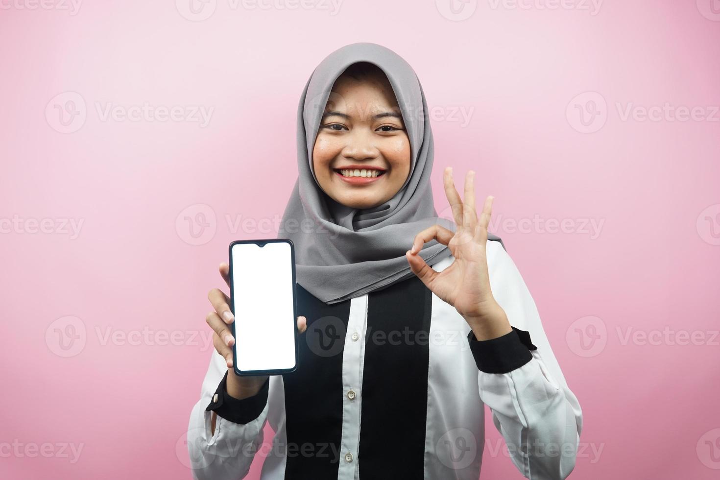 vacker ung asiatisk muslimsk kvinna som ler självsäkert och upprymt med händer som håller smartphone, presenterar applikation, ok tecken hand, bra jobb, framgång, isolerad på rosa bakgrund foto