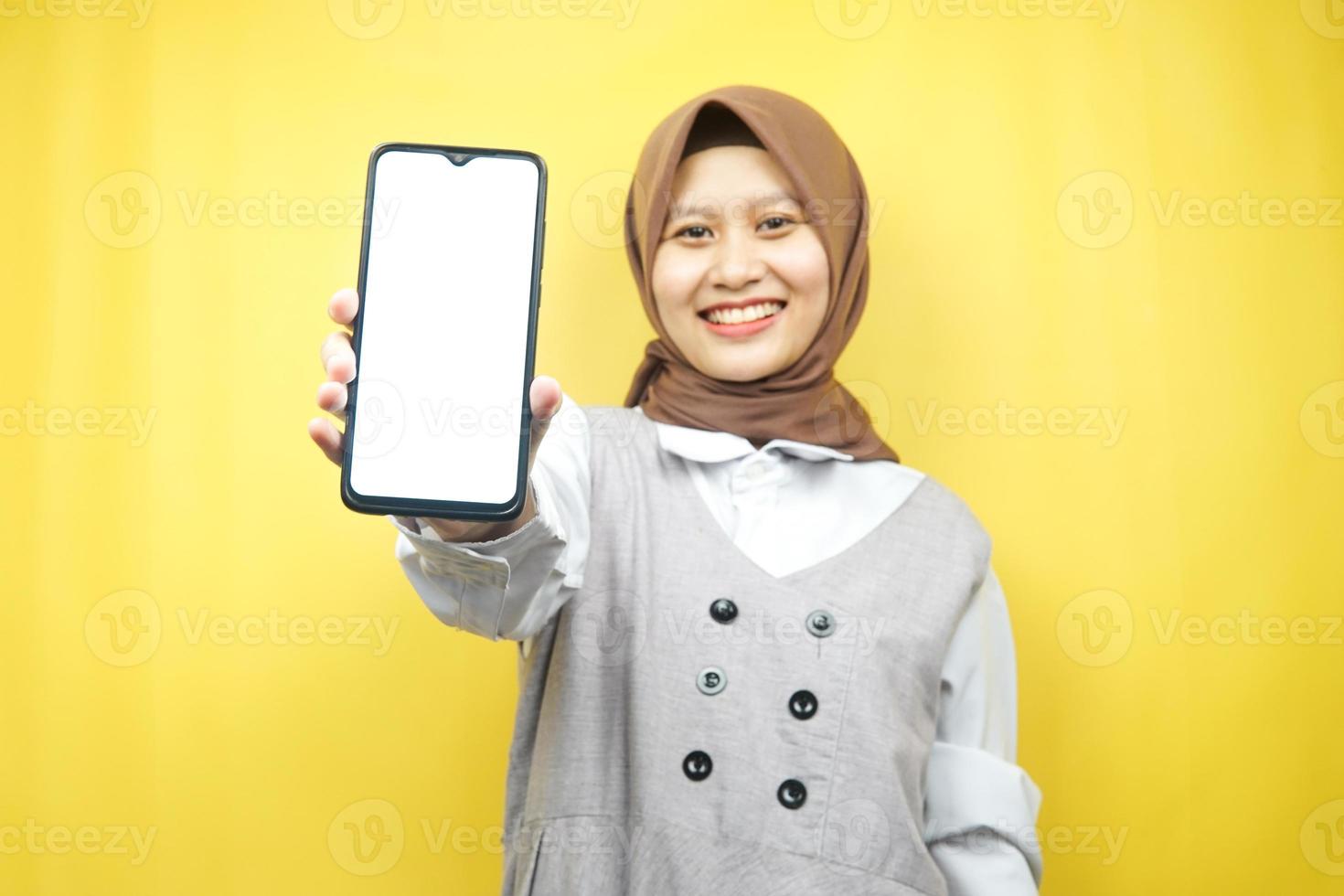 vacker ung asiatisk muslimsk kvinna som ler självsäker, entusiastisk och glad med händer som håller smartphone, främjar applikation, främjar något, isolerad på gul bakgrund foto