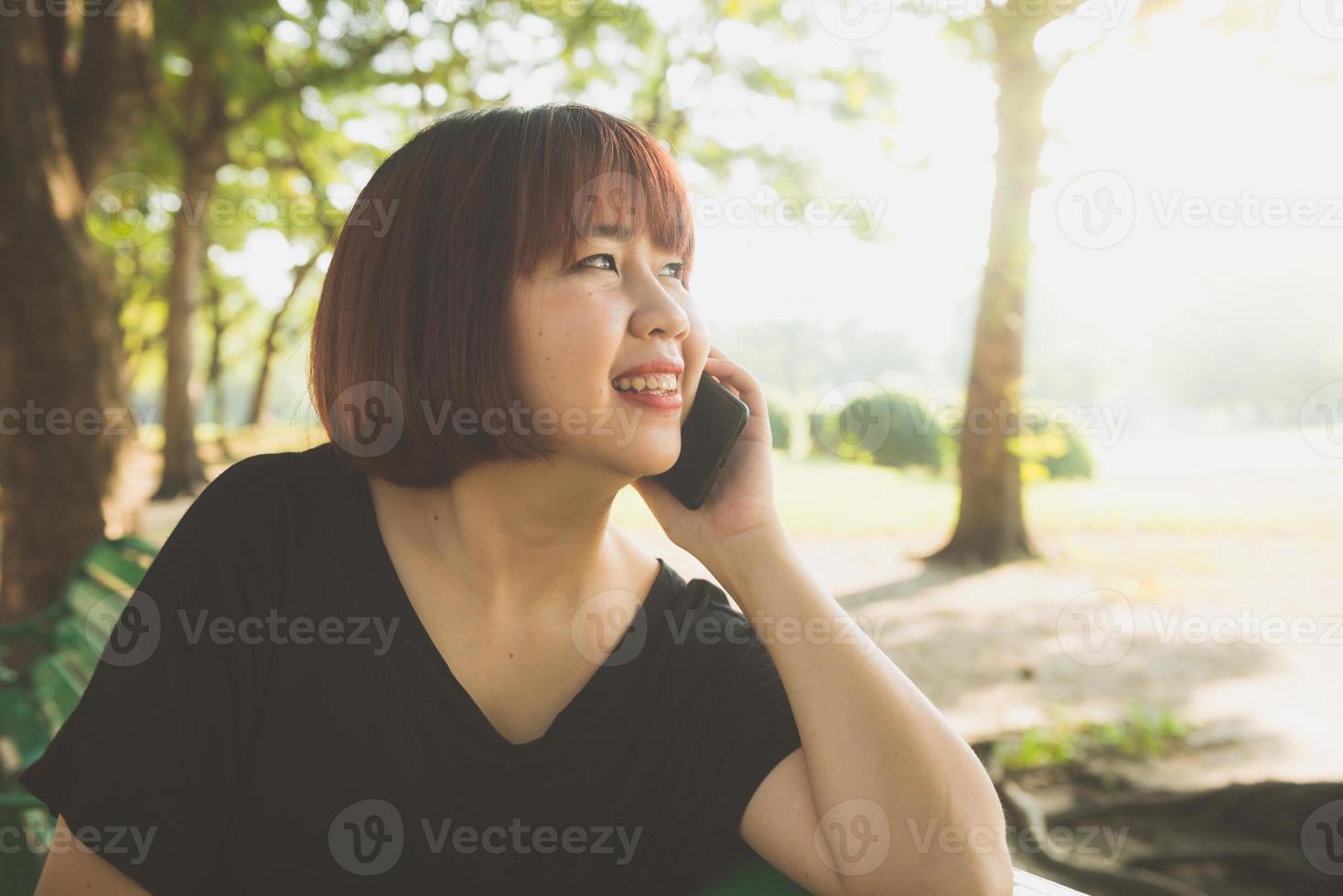 söt asiatisk kvinna ler och pratar i mobiltelefon medan du sitter i parken vårdag. asiatisk kvinna som använder på smart telefon med känsla av slappna av och smiley face. livsstil och teknikkoncept. foto