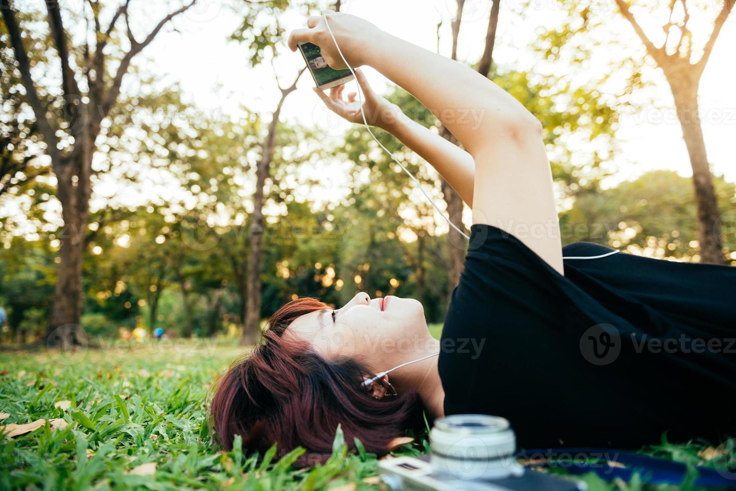 ung asiatisk kvinna som ligger på det gröna gräset och lyssnar på musik i parken med en chill känsla. ung kvinna avkopplande på gräset med sin kamera bredvid. utomhusaktivitet i parkkonceptet. foto