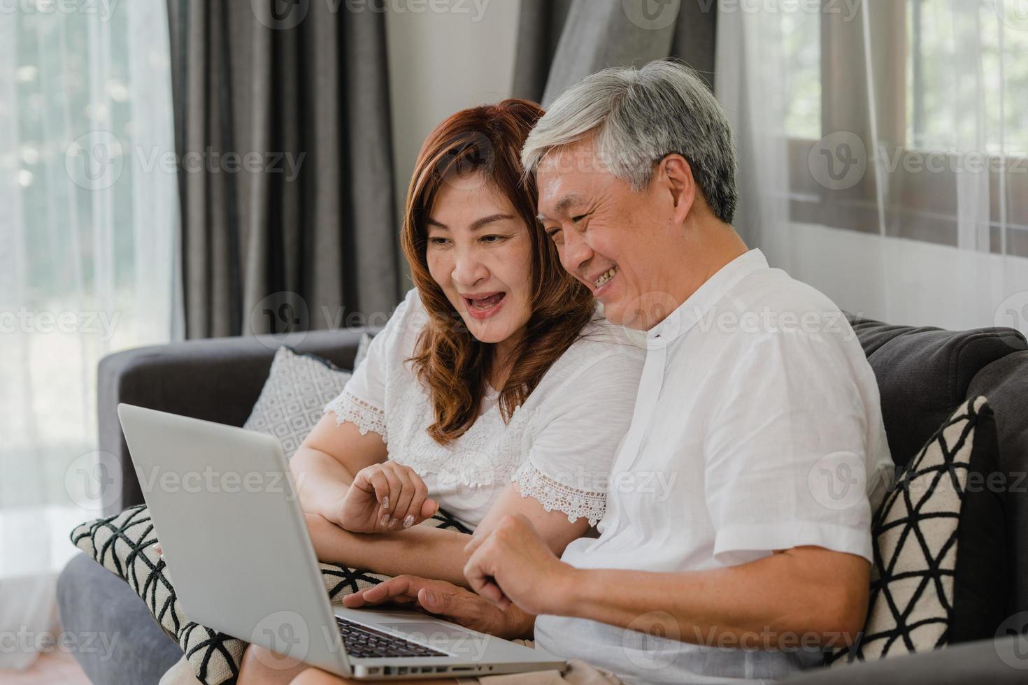 asiatiska senior par använder laptop hemma. asiatiska senior kinesiska morföräldrar, surfa på internet för att kolla sociala medier medan du ligger på soffan i vardagsrummet hemma koncept. foto