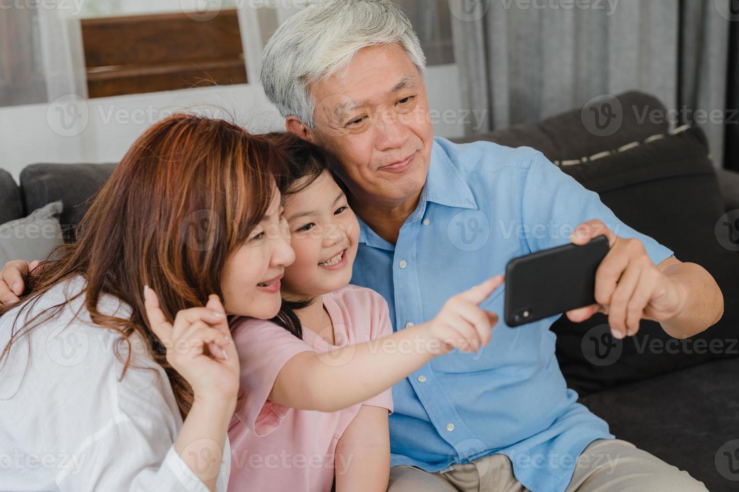 asiatiska morföräldrar selfie med barnbarn hemma. senior kineser, morfar och mormor tillbringar gärna familjetid koppla av med mobiltelefon med ung flickunge liggande på soffan i vardagsrumskonceptet. foto