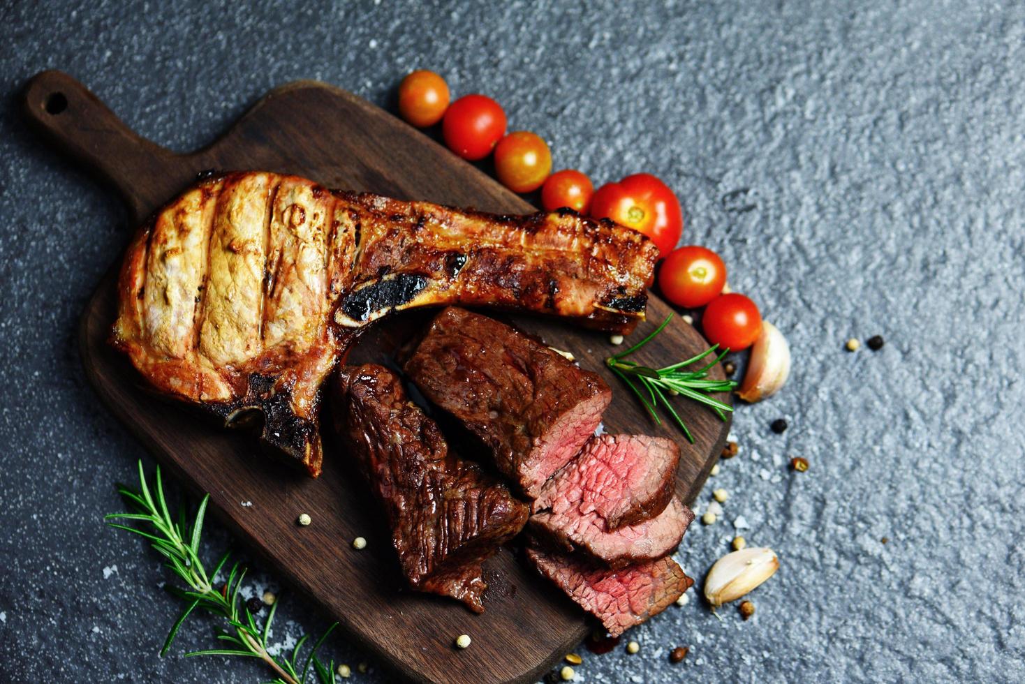 grillad nötköttskiva på svart bakgrund - rostad bifffilé och fläskkotletter med ört och kryddor servera med grönsaker på träskärbräda foto