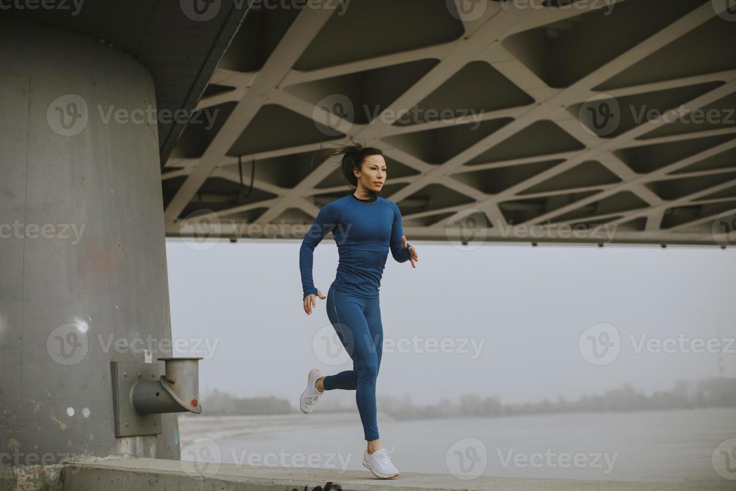 ung kvinna i blå träningsoverall springer vid floden på höstmorgonen foto