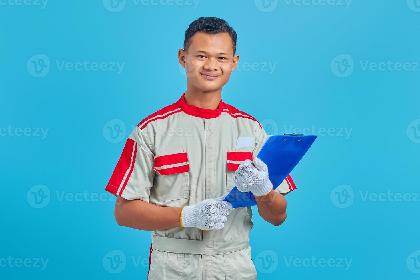 porträtt av glada unga asiatiska mekaniker håller urklipp isolerad på blå bakgrund foto