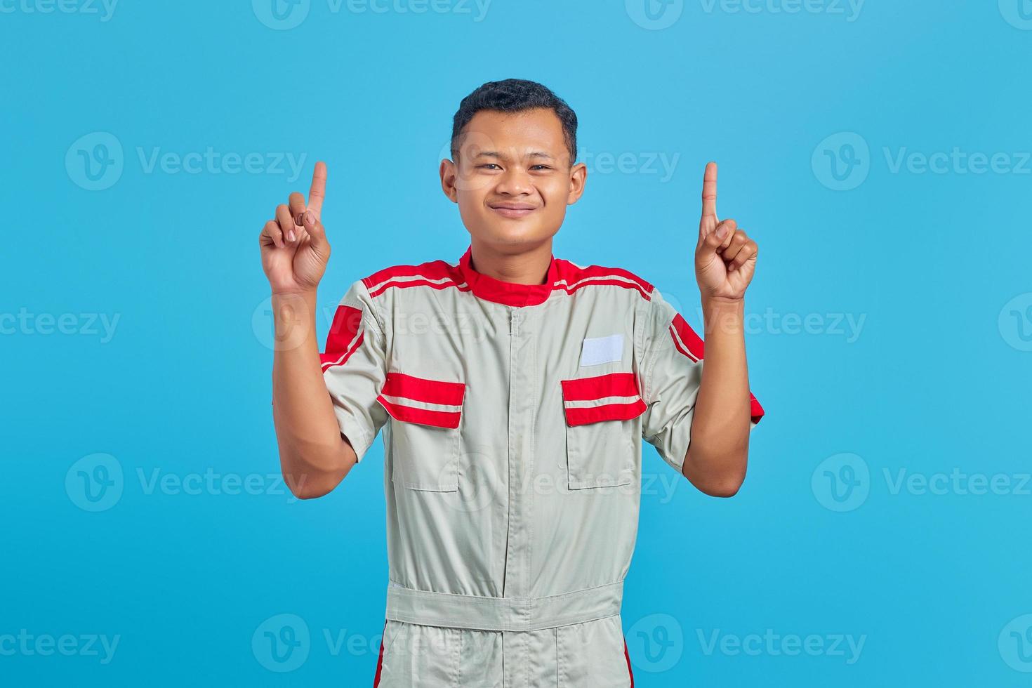 porträtt av leende ung asiatisk mekaniker medan lyft finger upp isolerad på blå bakgrund foto