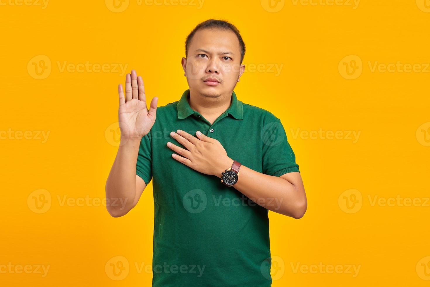 porträtt av stilig asiatisk man svär med händerna på bröstet och öppna handflator på gul bakgrund foto
