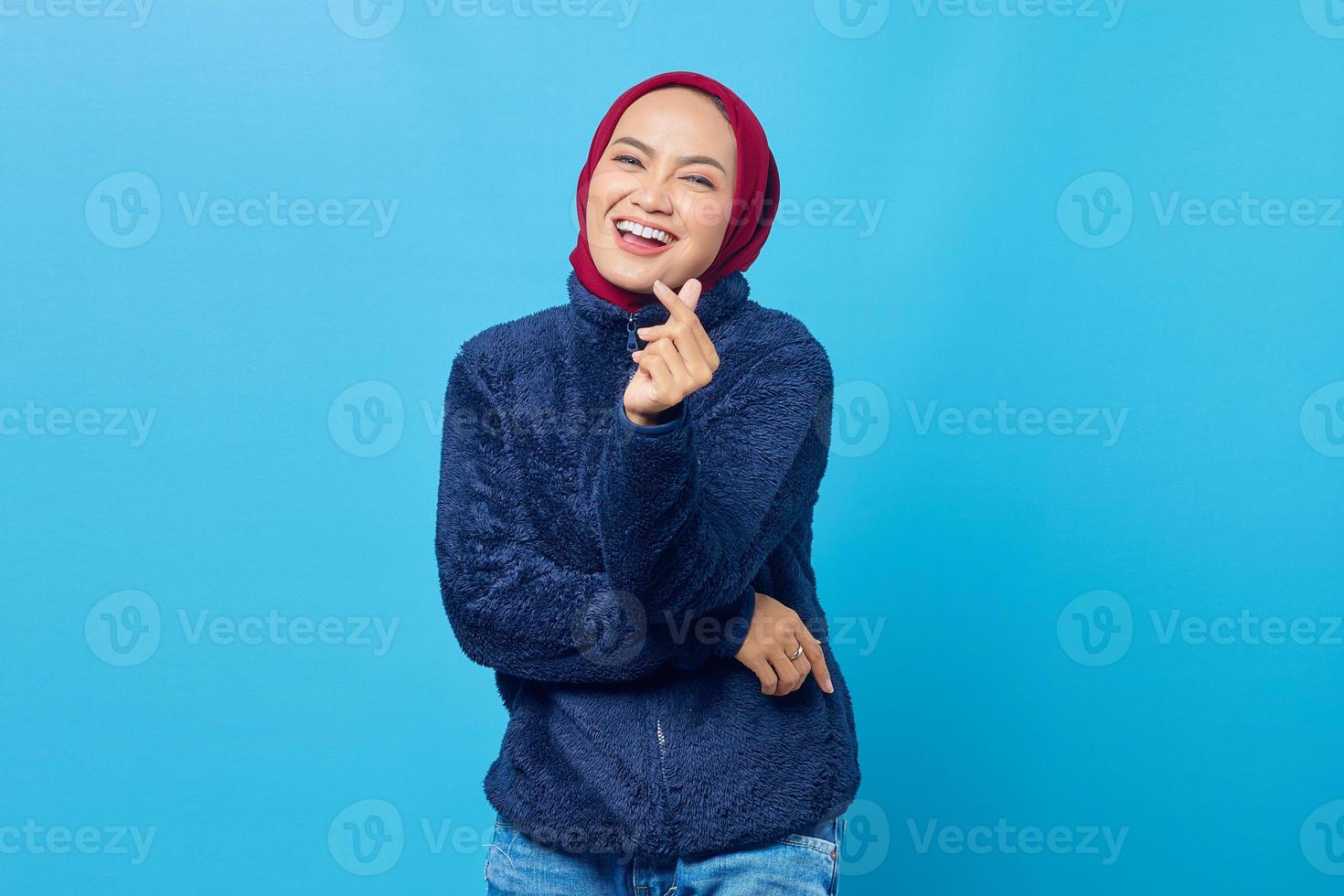 glad ung asiatisk kvinna visar hjärtat tecken med tummarna på blå bakgrund foto