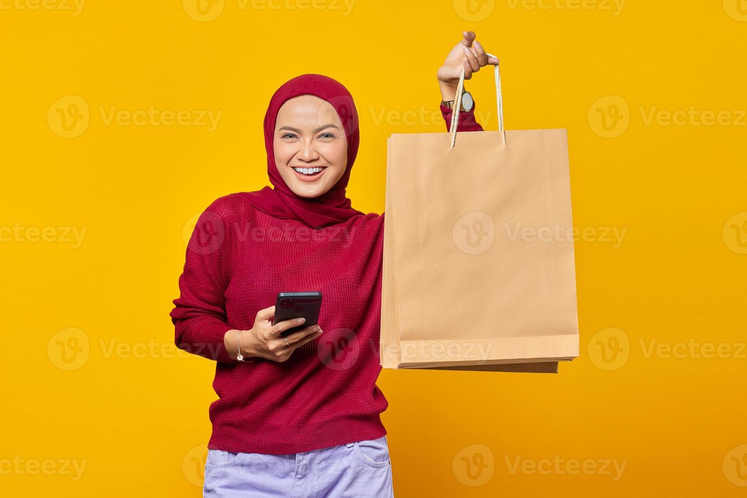 glad ung asiatisk kvinna som håller smartphone och visar shoppingkassar på gul bakgrund foto