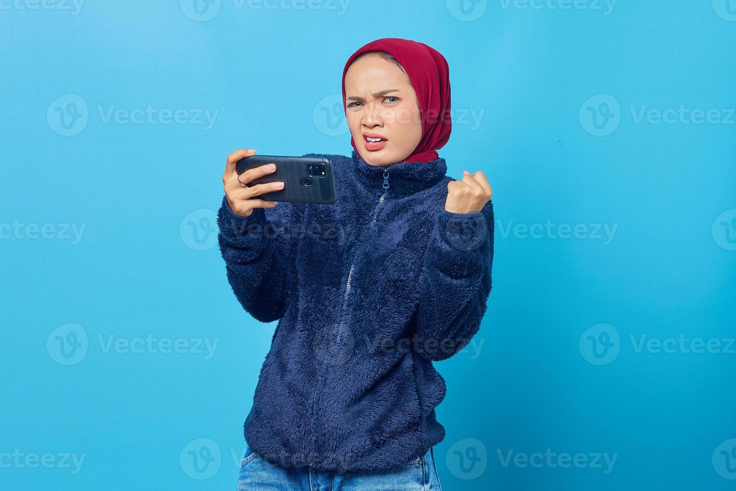 ledsen ung vacker kvinna spelar ett spel på mobiltelefon på blå bakgrund foto