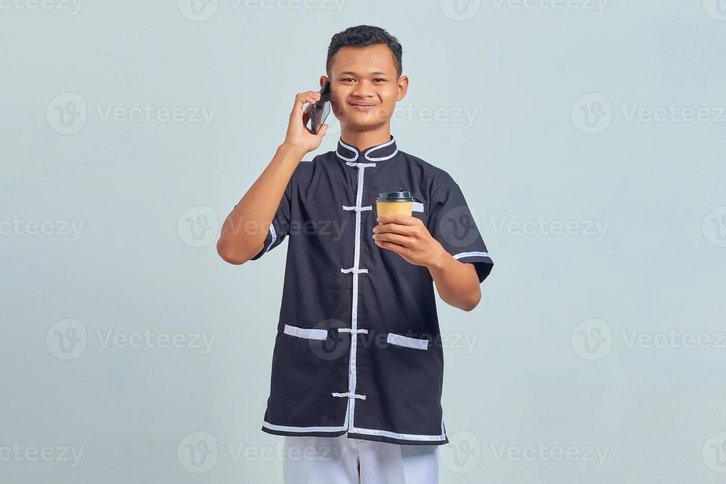 porträtt av leende asiatisk ung man iklädd karateuniform som pratar på smartphone och håller kaffe på grå bakgrund foto