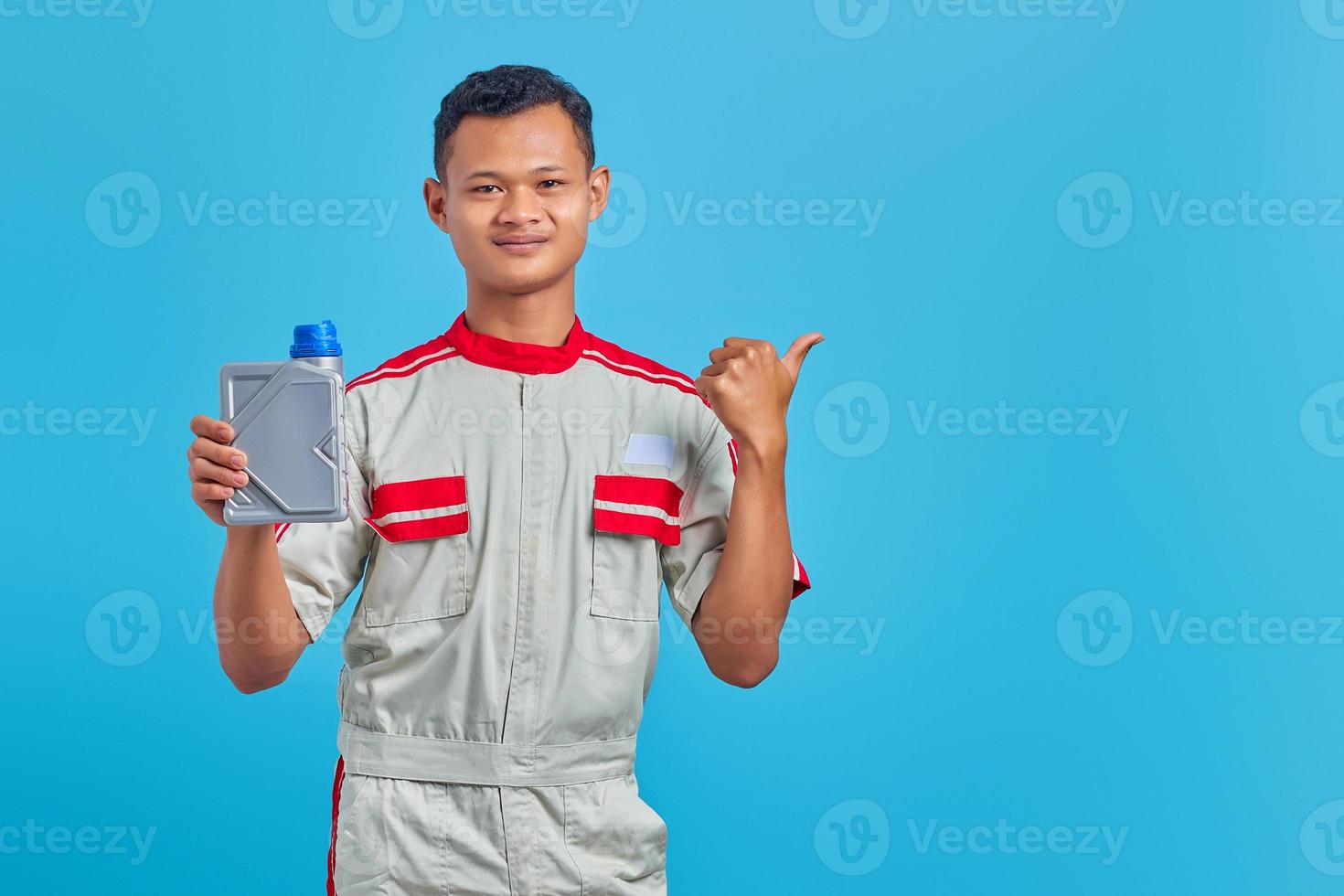 porträtt av leende ung asiatisk mekaniker som visar motorolja plastflaska och pekar på tomt utrymme över blå bakgrund foto