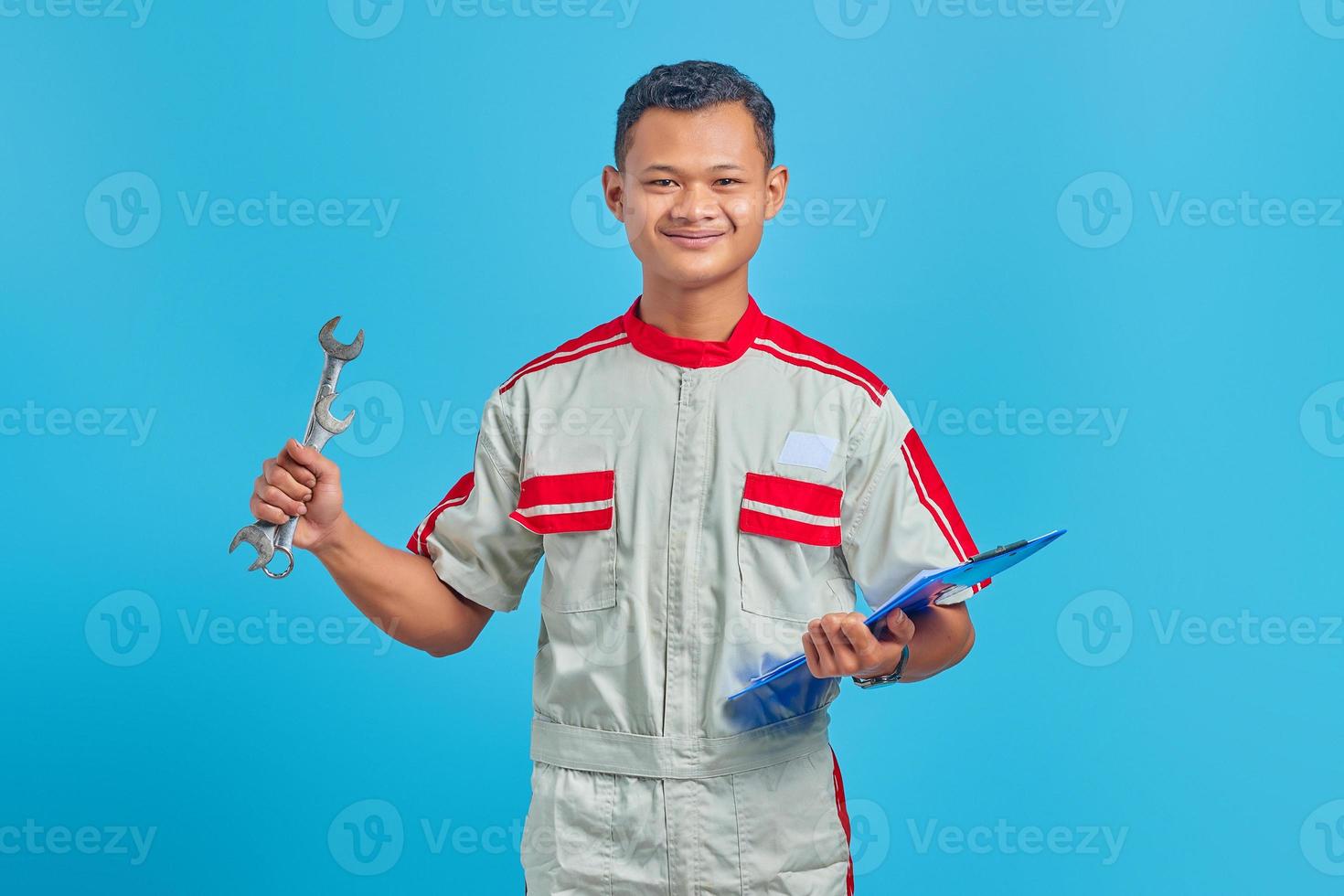 porträtt av en leende ung mekaniker som håller i urklippet och visar skiftnyckel isolerad på blå bakgrund foto