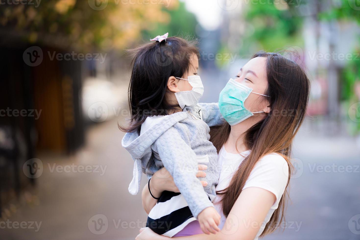 koncept mamma som tar hand om dotter bär medicinsk ansiktsmask. mamma bar och omfamnade barnet med kärlek på allmän väg. båda bär mask för att skydda mot damm och virus. foto