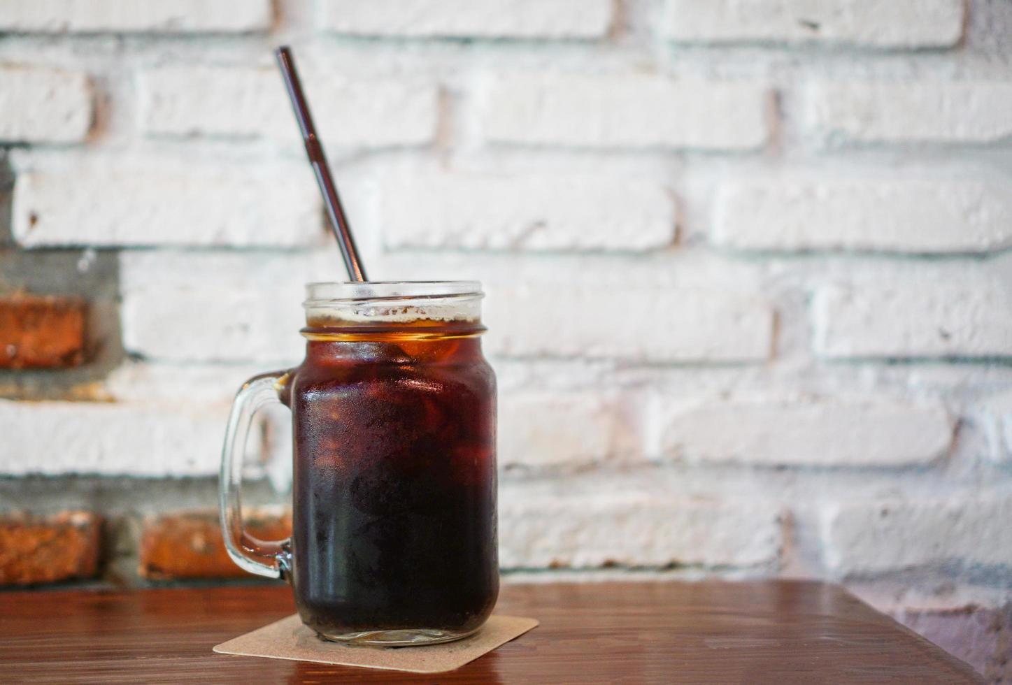 glas iced svart kaffe i burk på trä bordet med vägg tegel bakgrund foto