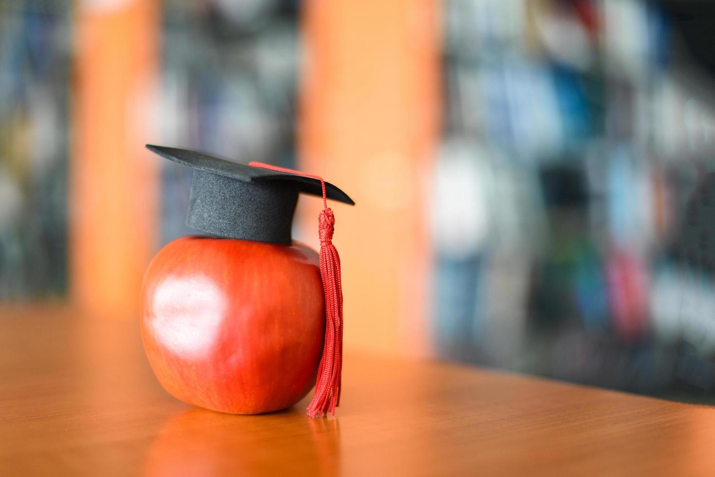 utbildning lärande koncept - examen mössa på äpple på bordet med bokhylla i biblioteket bakgrund foto