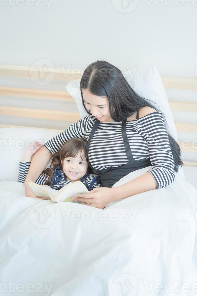 lycklig kärleksfull familj ung asiatisk mamma läser rolig bok med sin söta dotter i sovrummet. fotodesign för familj, barn och glada människor koncept foto