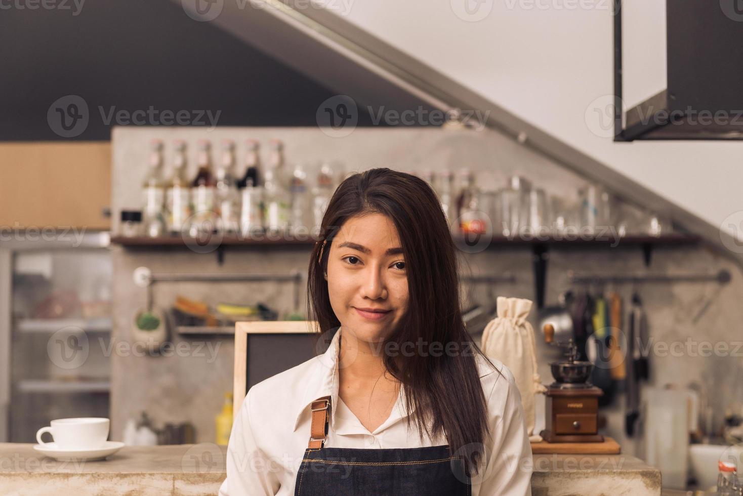 protrait av ung asiatisk kvinnlig barista välkomnar sin kund till kafé i en varm ljus eftermiddag med ett vackert leende. ung kvinnlig barista leende i hennes lilla kafé. mat och dryck koncept. foto
