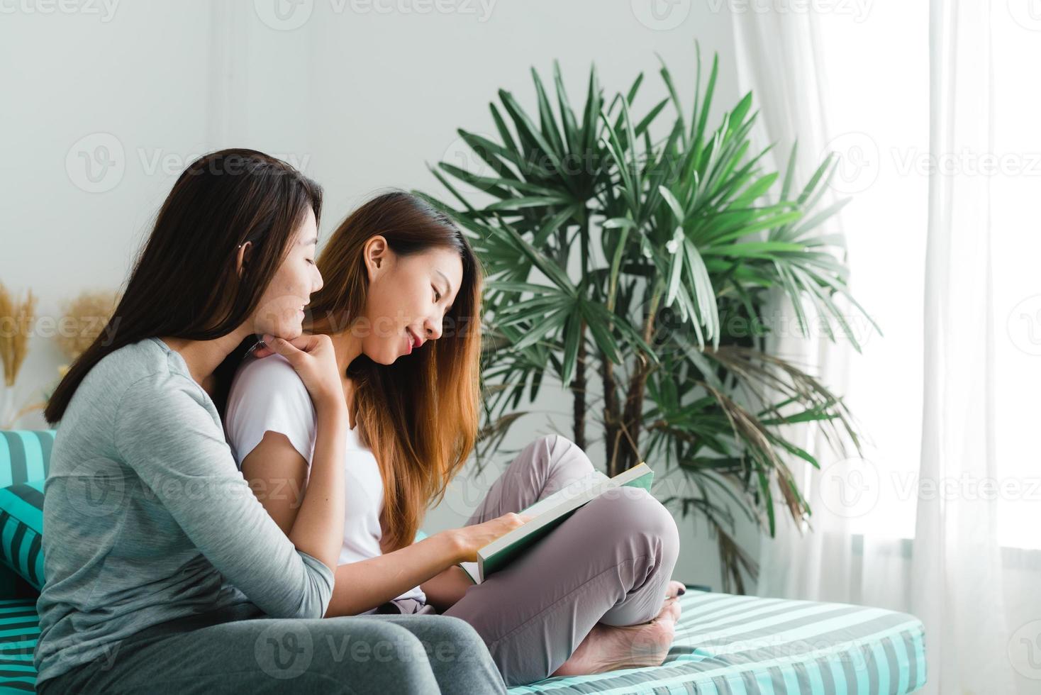 vackra unga asiatiska kvinnor hbt lesbisk lyckligt par sitter på sängen och läser bok tillsammans nära fönstret i sovrummet hemma. hbt lesbisk par tillsammans inomhus koncept. spendera trevlig tid hemma. foto
