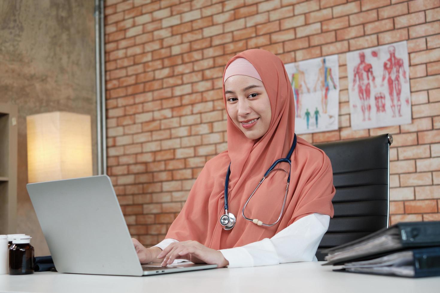 porträtt av en vacker kvinnlig läkare, vacker muslim i uniform med ett stetoskop, ler och arbetar med laptop på sjukhusets klinik. en person som har expertis inom professionell behandling. foto