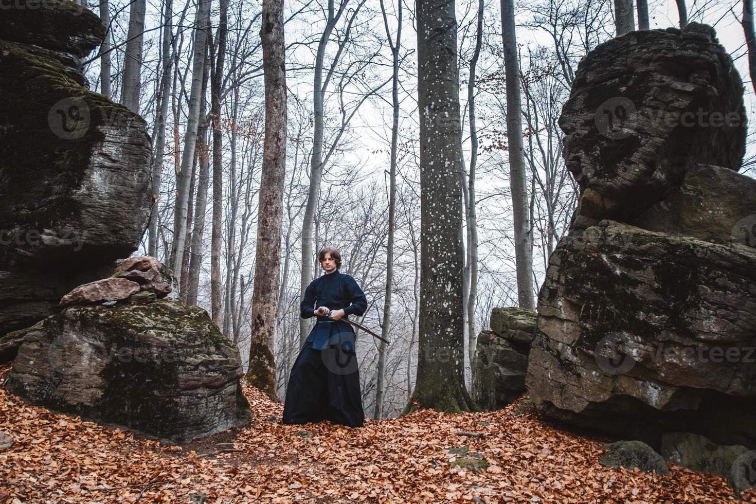 man i svart kimono utövar kampsport med ett svärd på stenar och skog bakgrund foto