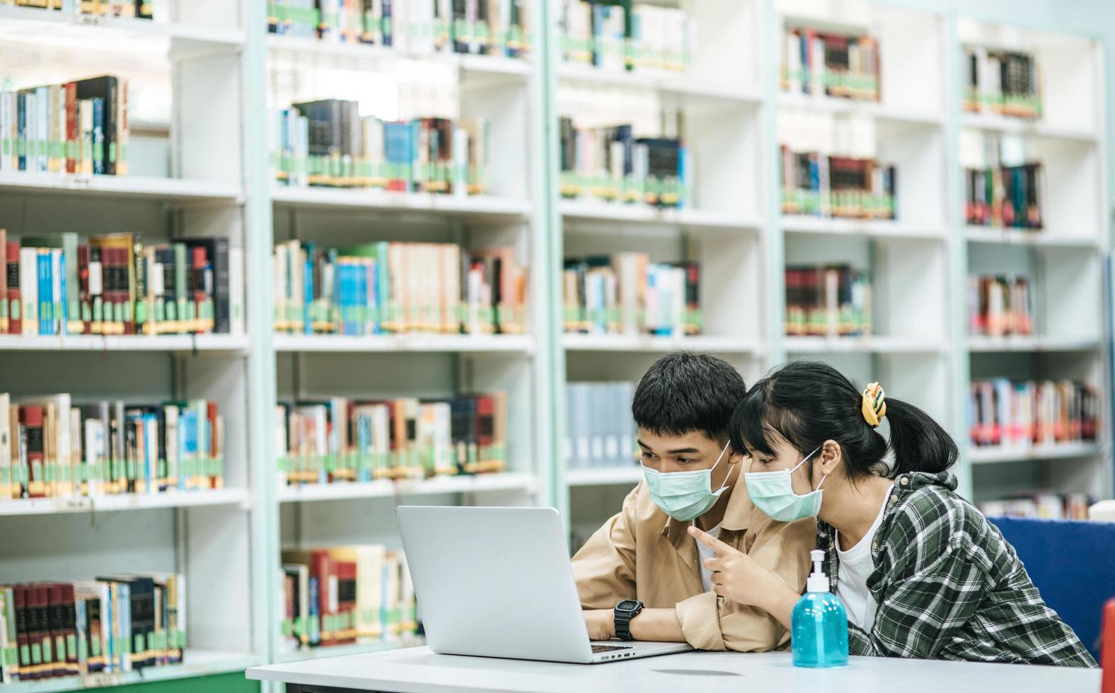 män och kvinnor bär masker och använder en bärbar dator för att söka efter böcker i biblioteket. foto