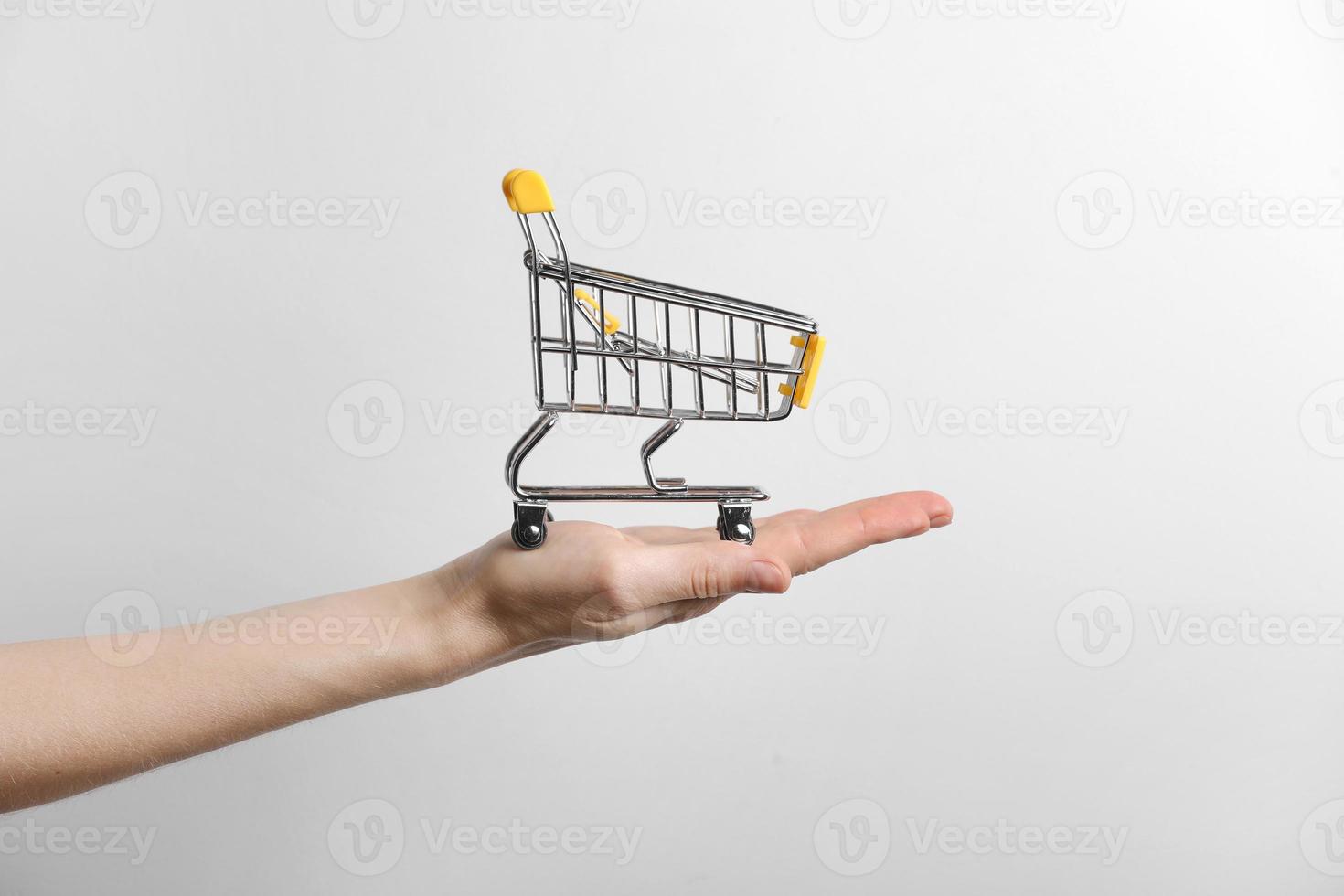närbild av en kvinnas hand som håller en liten tom metallvagn i handflatan på grå bakgrund. konceptet shopping. kopieringsutrymme för reklam. foto